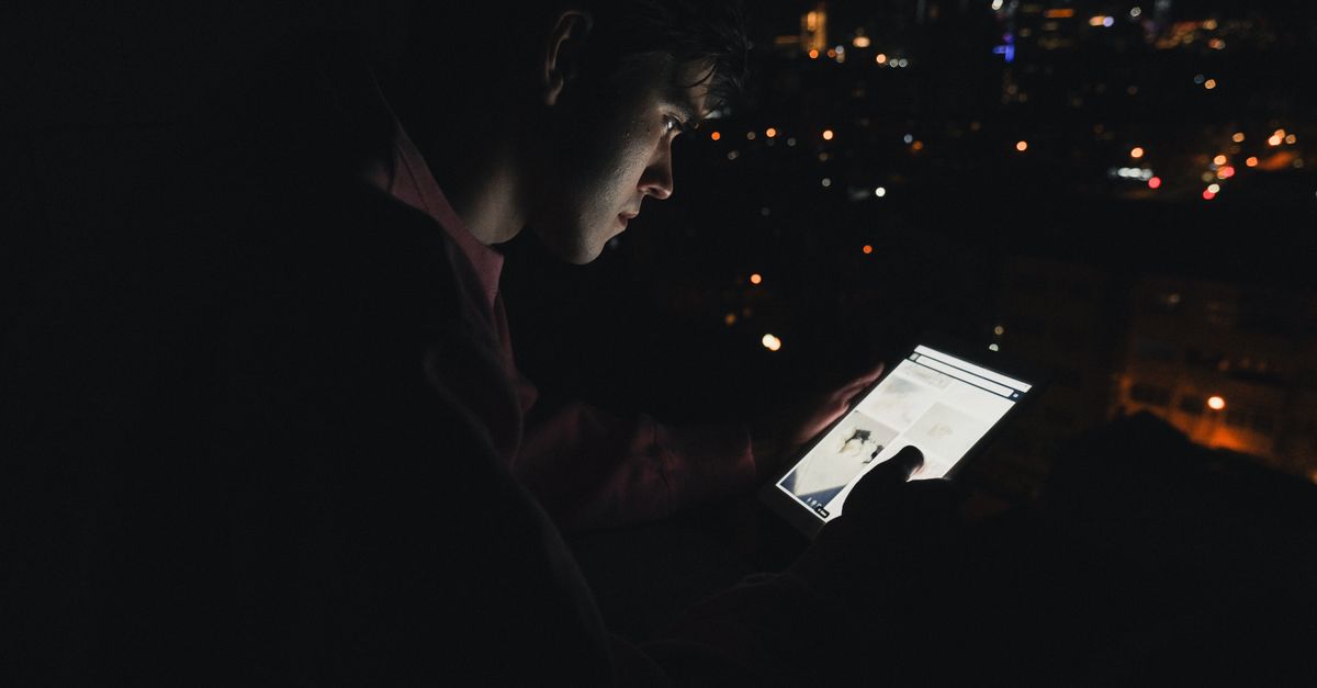 Silhouette of a man using a glowing digital tablet with city lights in the background at night.